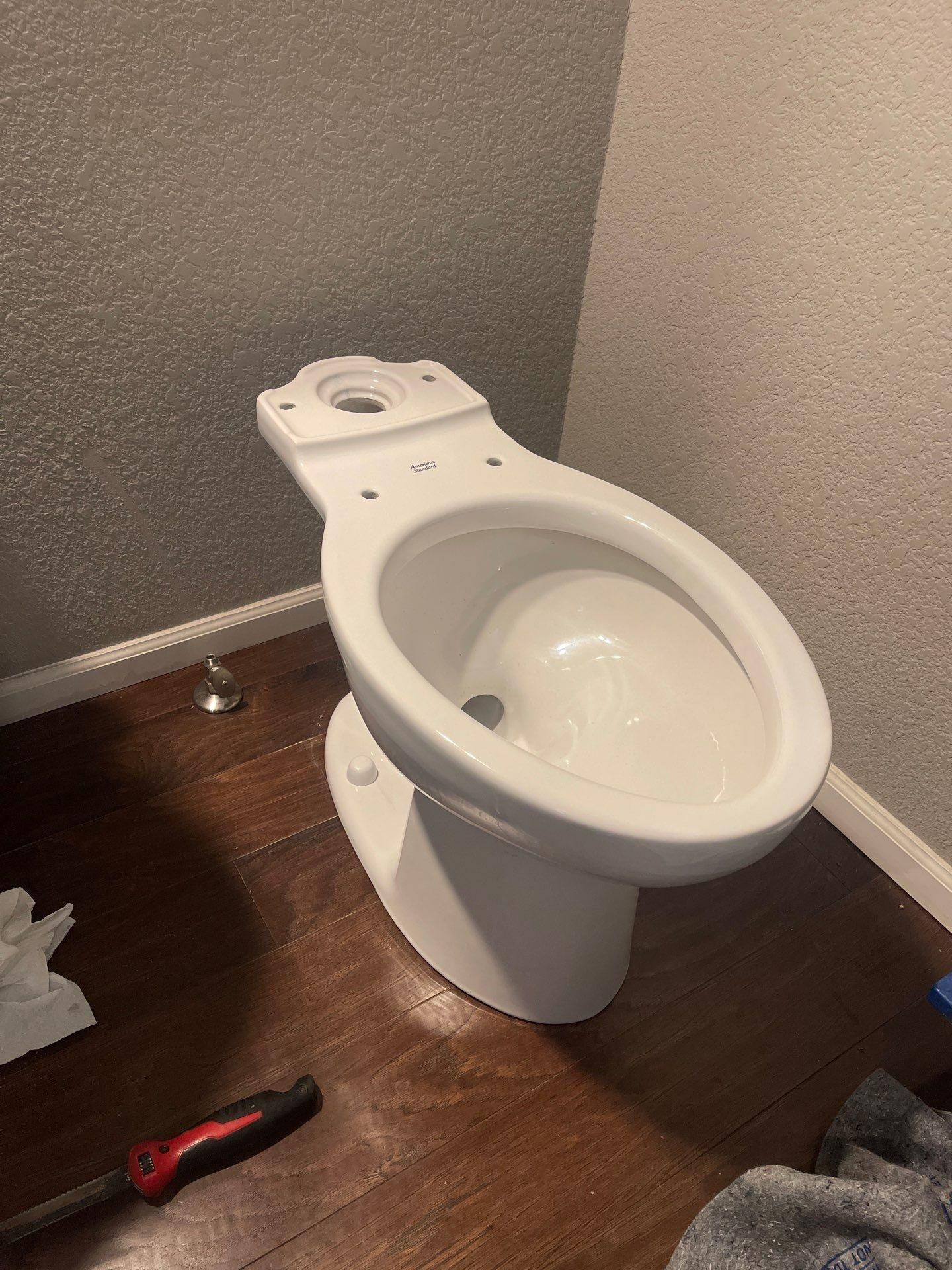 Toilet in a corner on a wood floor, with a red screwdriver nearby.
