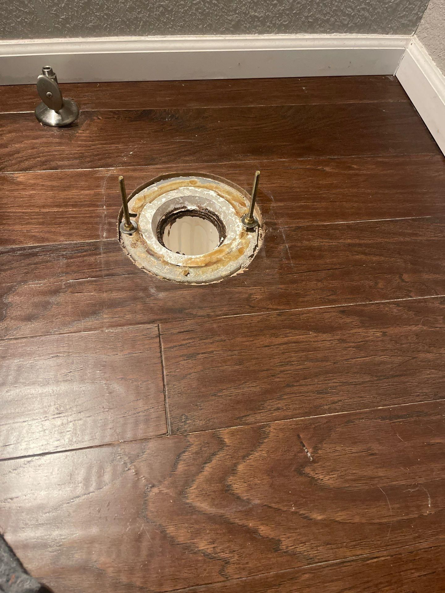 Damaged hardwood floor with a circular opening and exposed pipe fitting near the baseboard