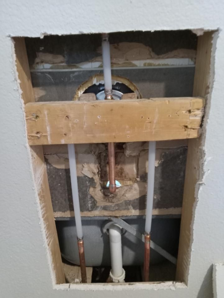Opened wall with exposed plumbing pipes and copper lines around a damaged hole and wooden brace.