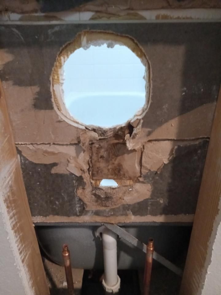 Damaged wall with large circular hole and exposed plumbing beneath, viewed from inside.