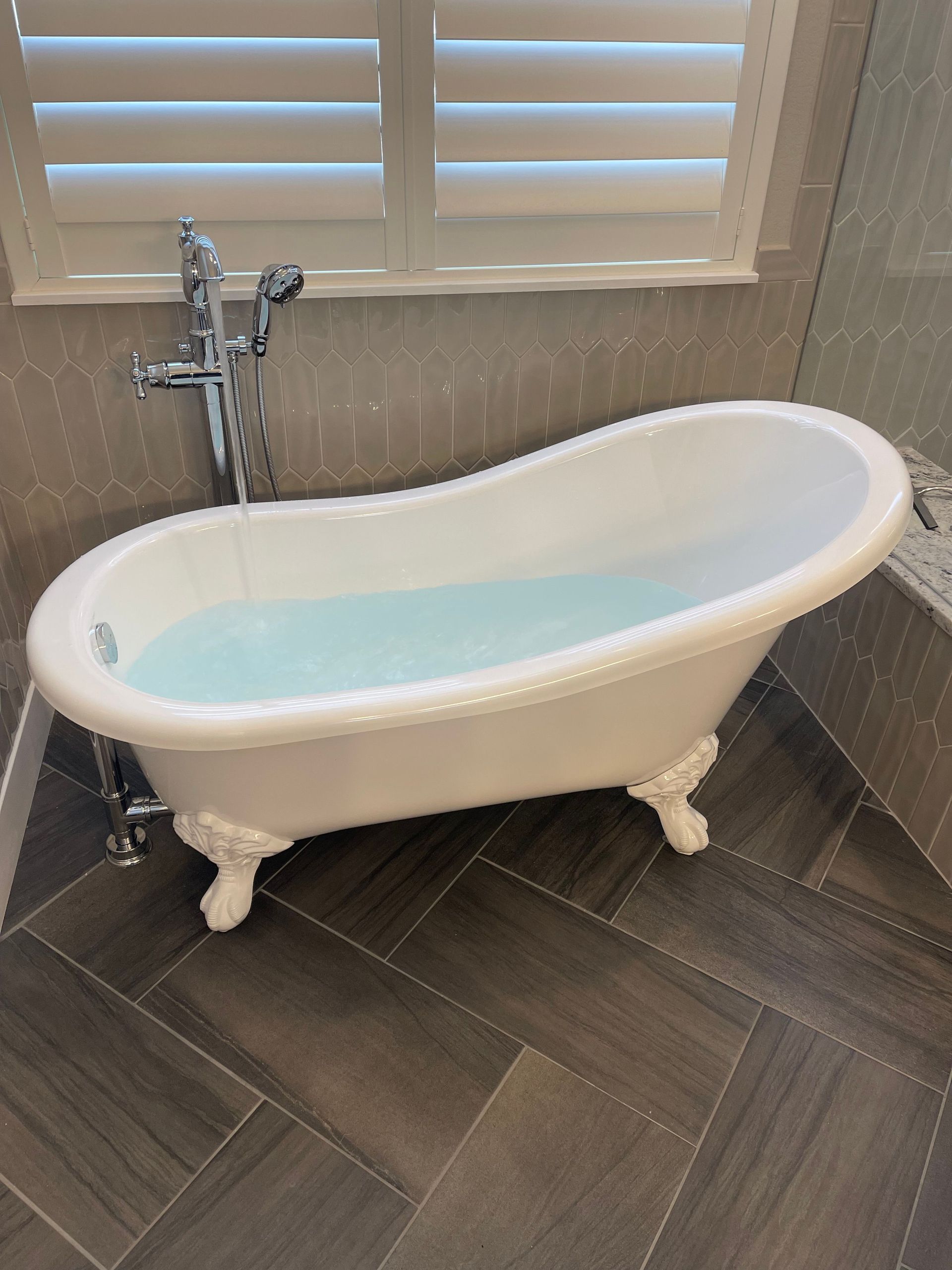 Clawfoot bathtub filled with water in a tiled bathroom beside a window with shutters