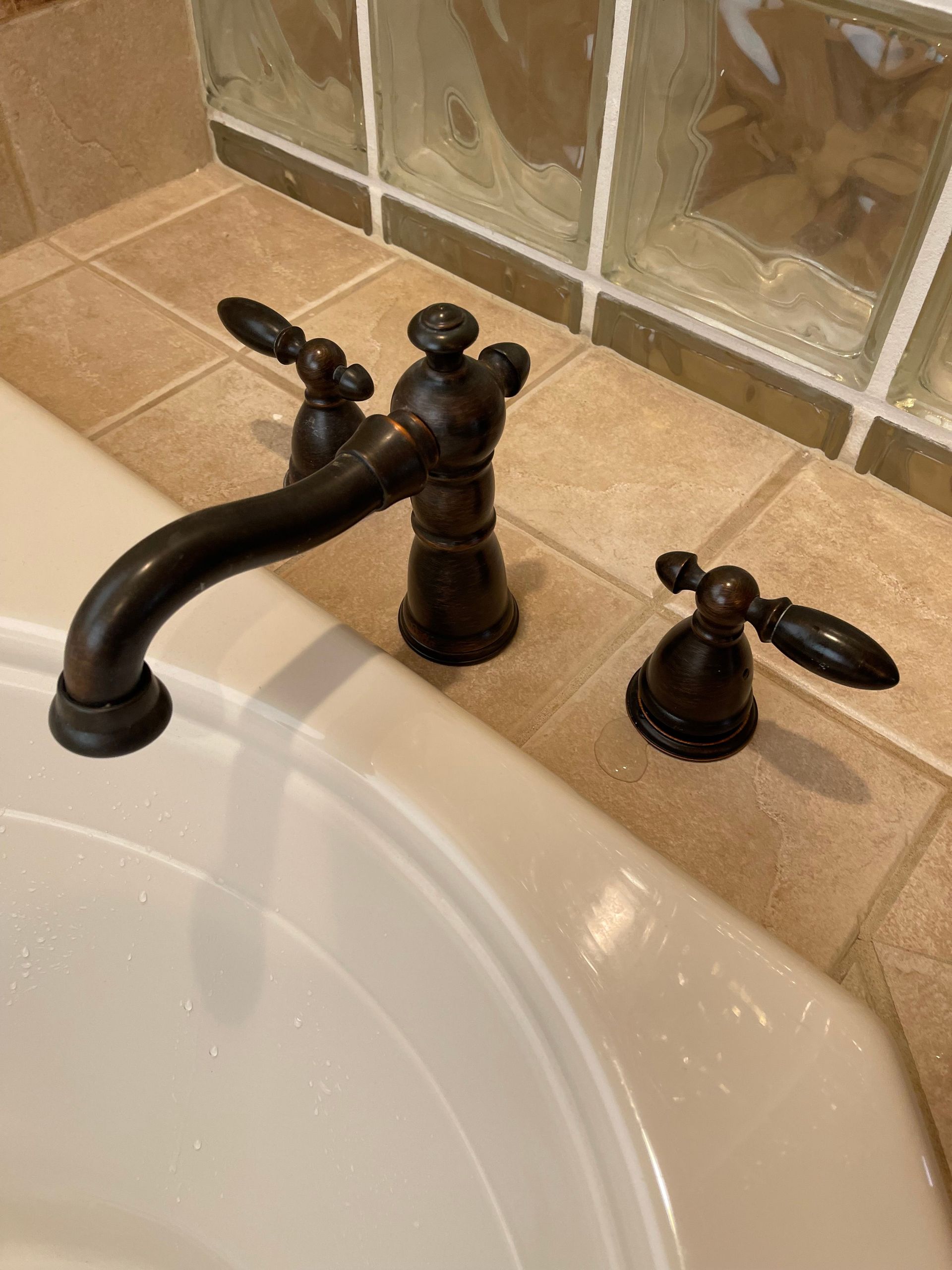 Bathroom sink with dark bronze faucet and handles on beige countertop, beside a tiled backsplash.