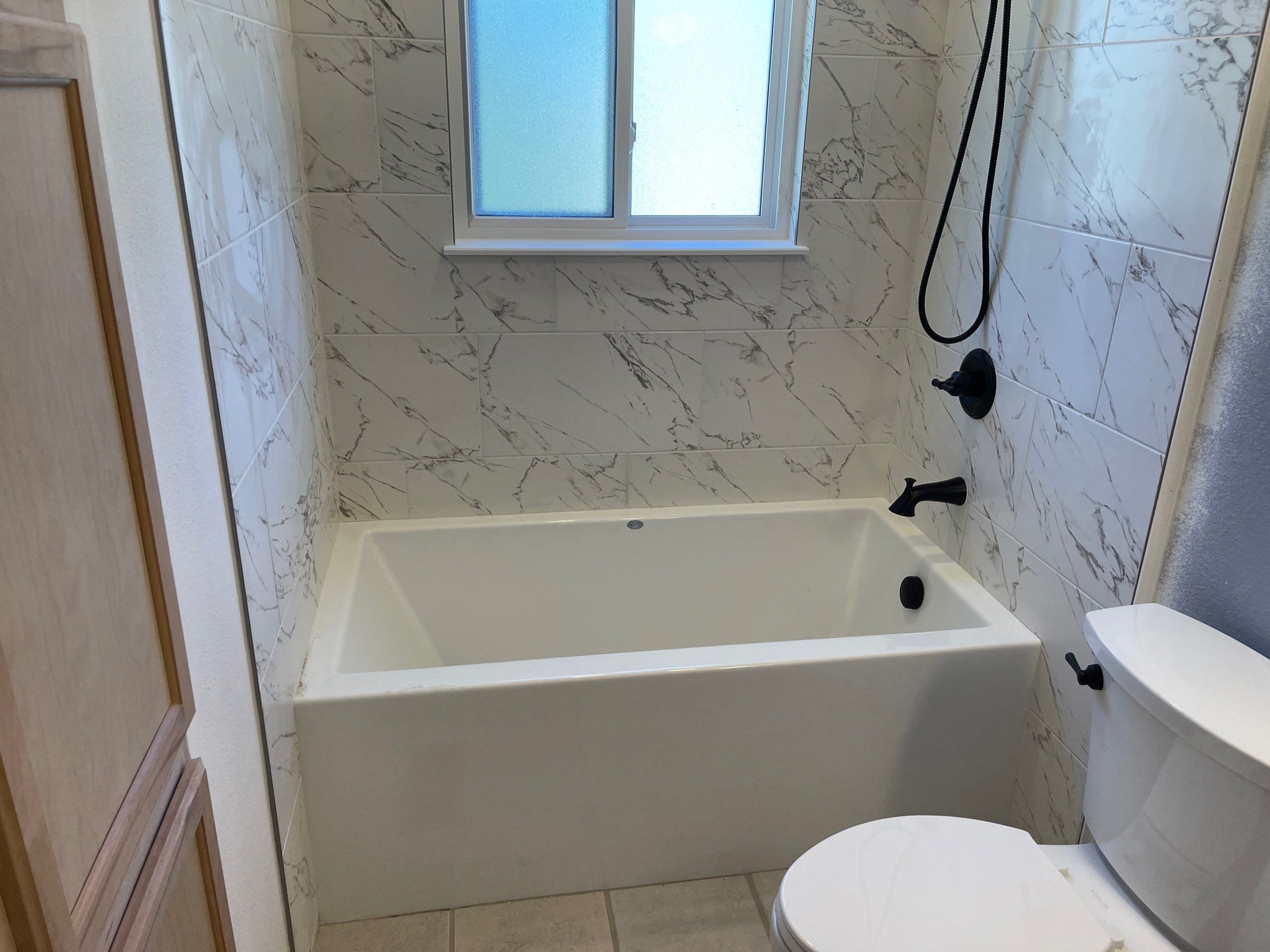 Small bathroom with a white bathtub, marble-look walls, window, and toilet.