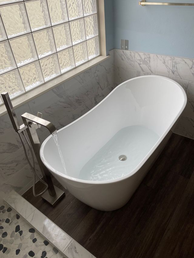 Modern white freestanding bathtub beside a frosted window in a tiled bathroom