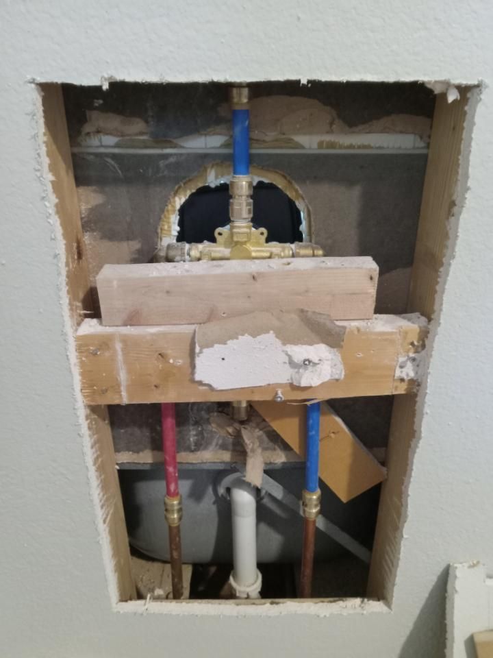Open wall with exposed plumbing pipes and rough drywall repair around a bathroom fixture opening