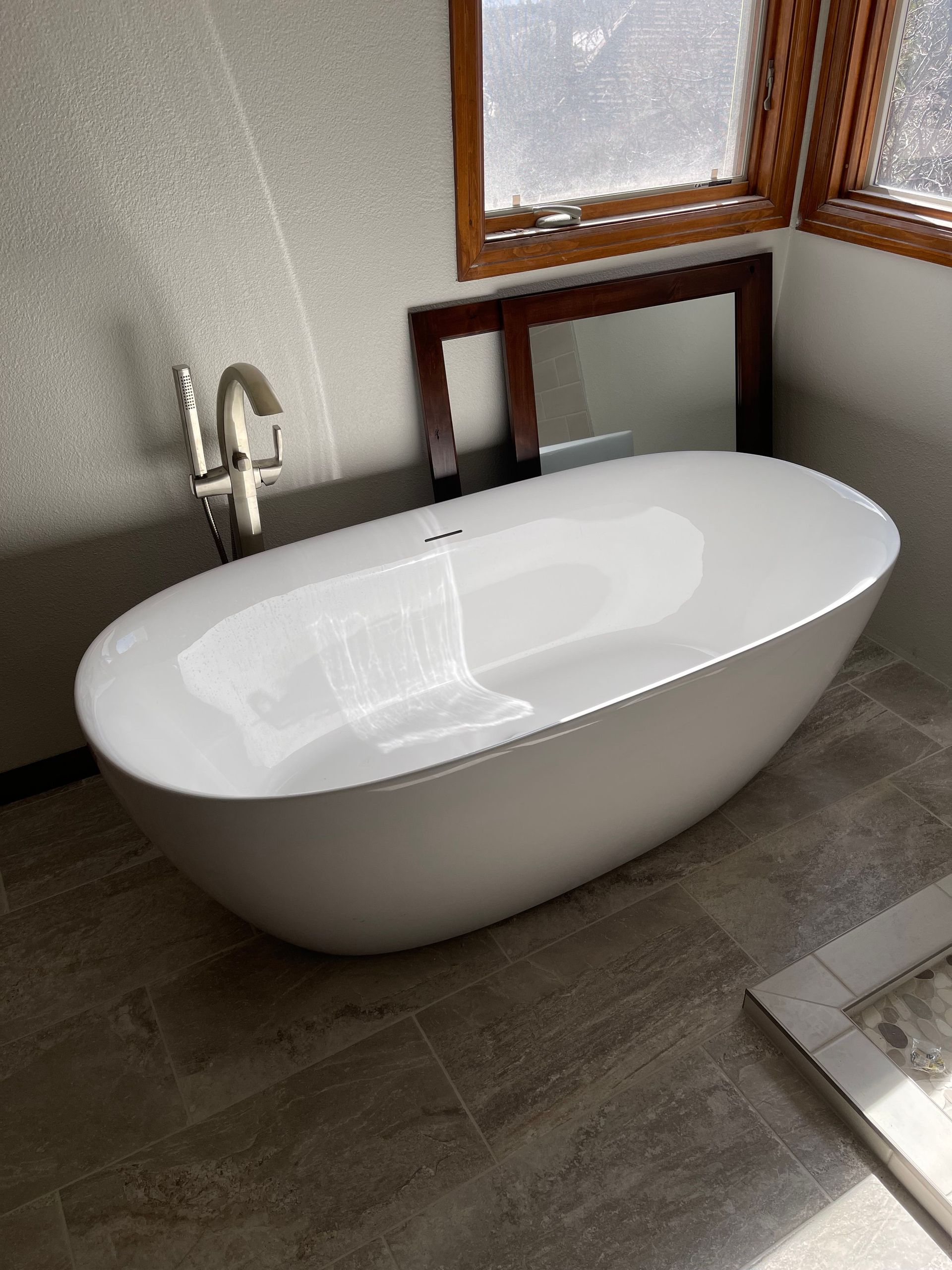 Modern white freestanding bathtub in a bathroom with wood-framed windows and gray tile floor