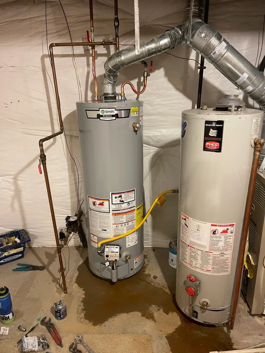 Two water heaters in a basement with pipes and a wet floor beneath them