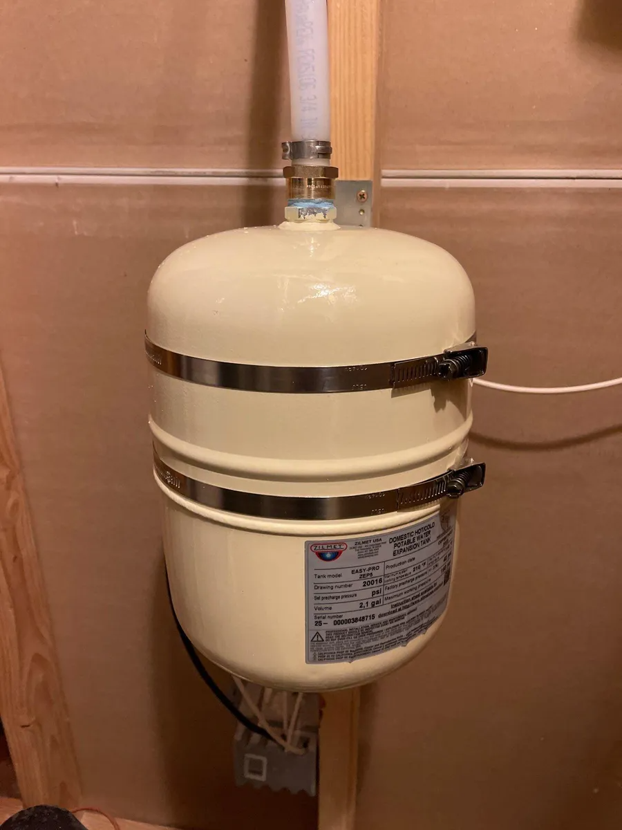Cream-colored wall-mounted expansion tank with label and connected pipes on a beige wall.