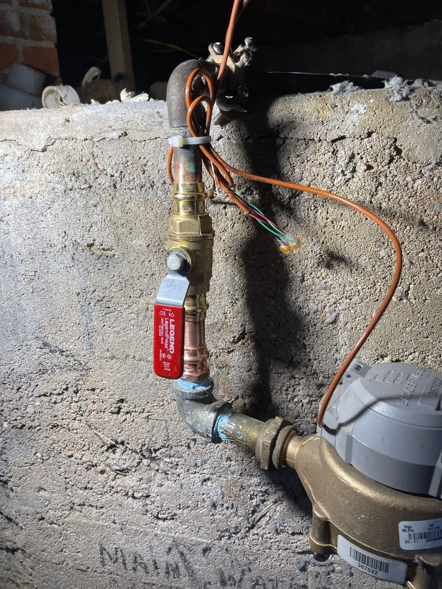 Water meter and shutoff valve on a rough concrete wall, with exposed orange wire and piping.