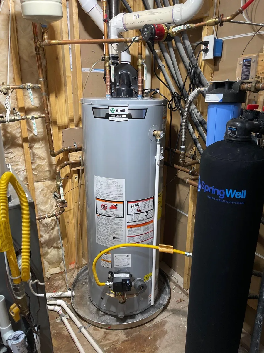 Water heater in a utility room, with exposed pipes, yellow hoses, and a black water softener tank nearby
