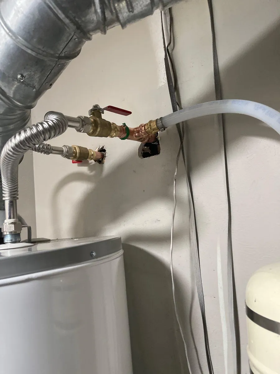 Water heater plumbing with metal and white hoses, valves, and ducting in a utility closet.