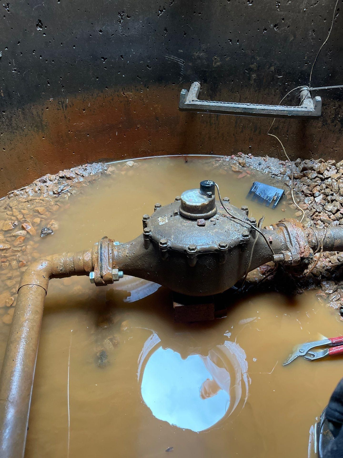 Rusty pipe valve in a muddy pit filled with brown water, with a small blue object nearby