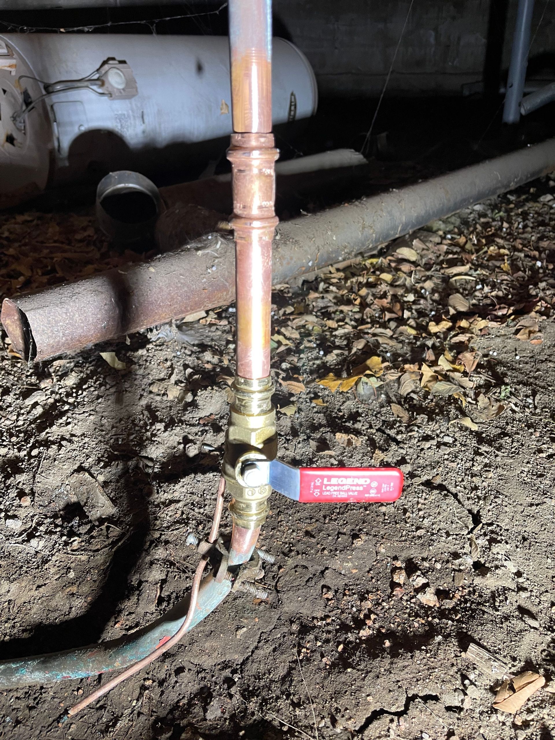 Close-up of a red-handled valve on a copper pipe over a dirty basement floor.