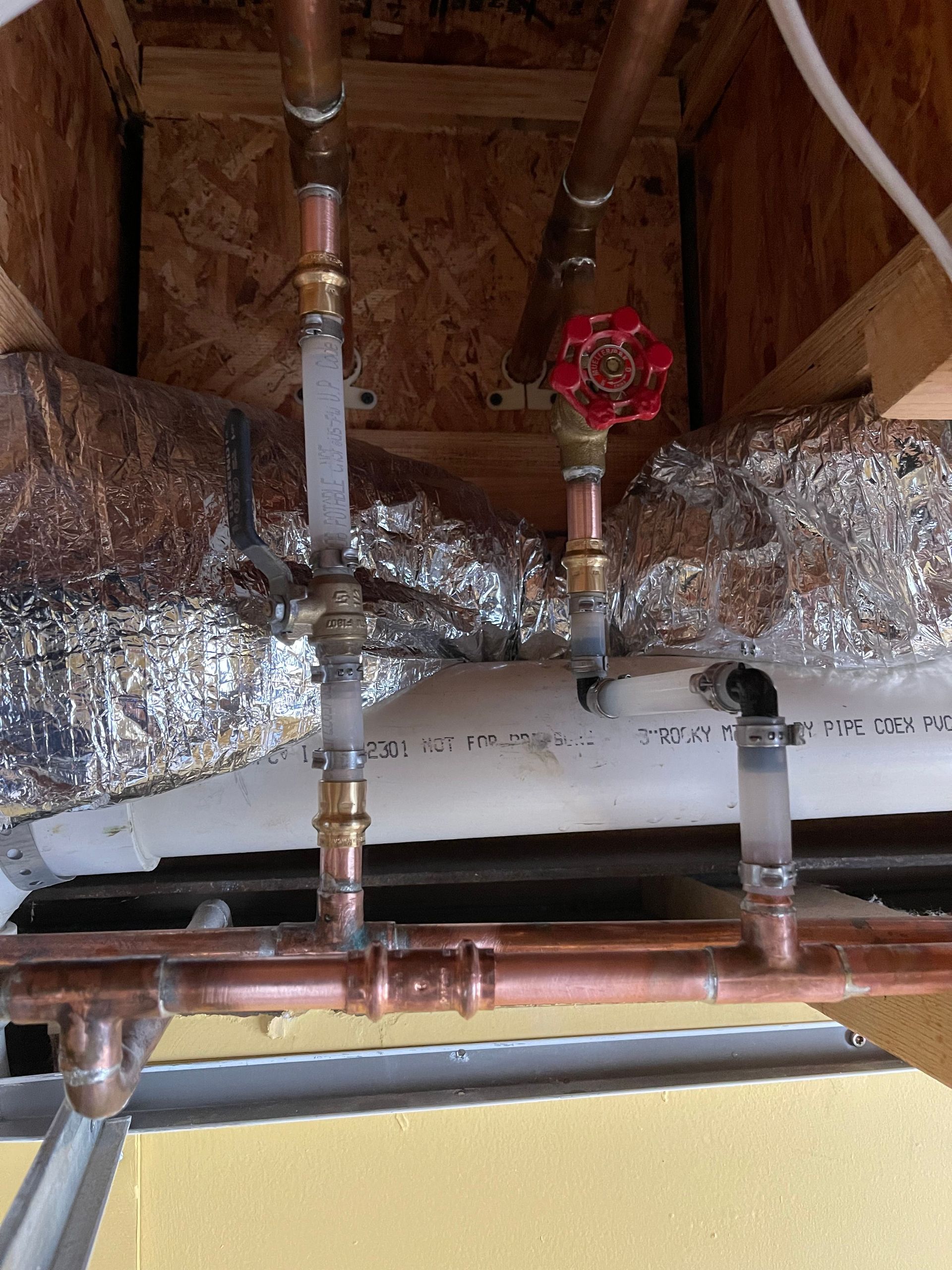 Plumbing pipes and valves under a house, with insulated ducts and exposed wooden joists.