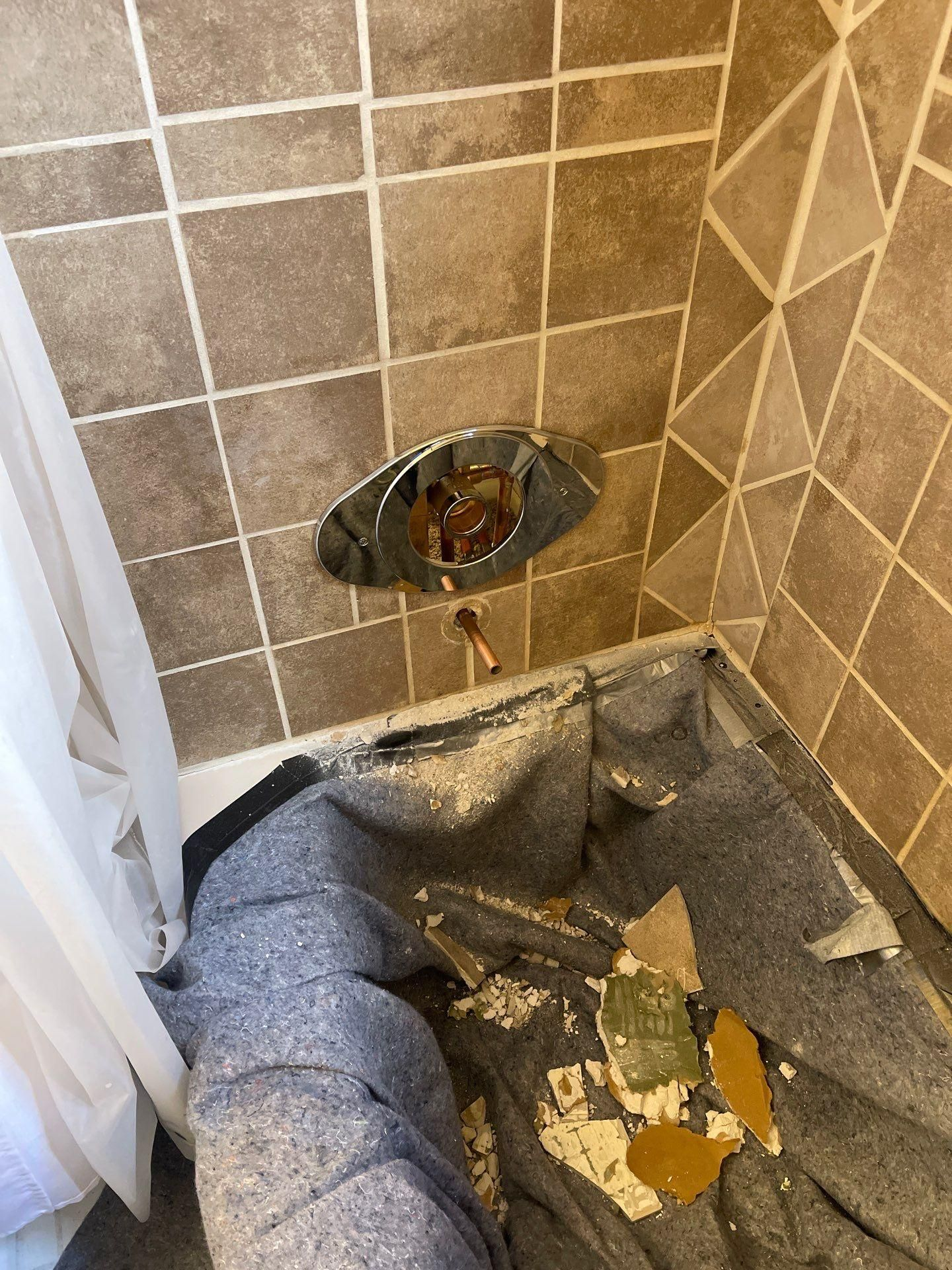 Damaged bathroom wall with exposed plumbing and debris on the shower floor