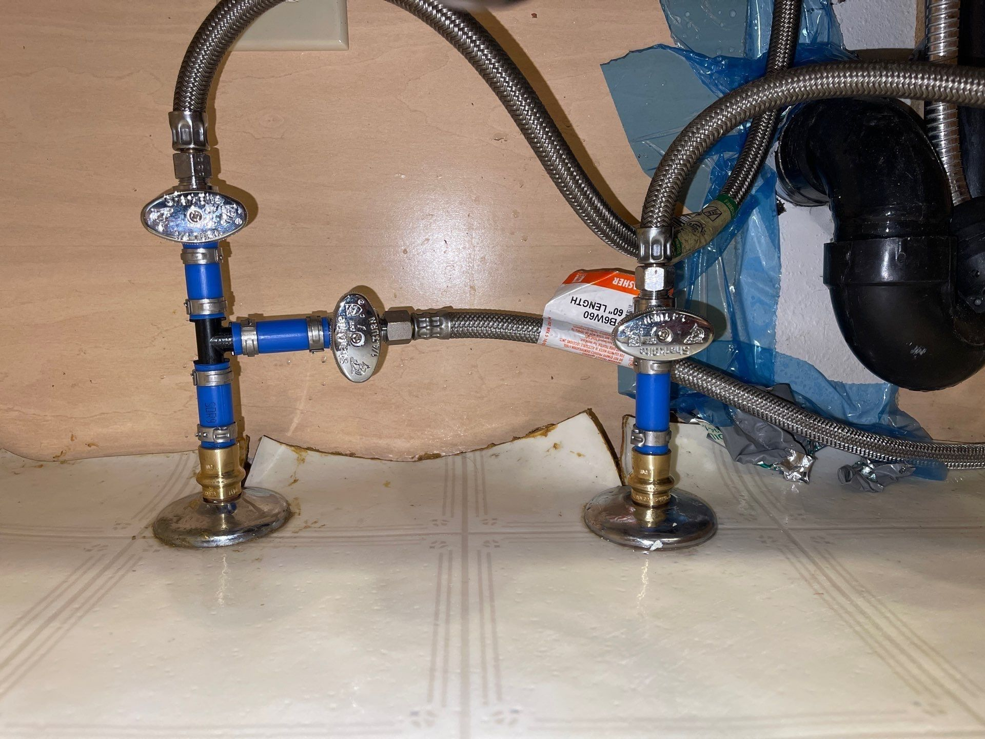 Under-sink plumbing with blue and chrome pipes connected to two shutoff valves on a tiled floor
