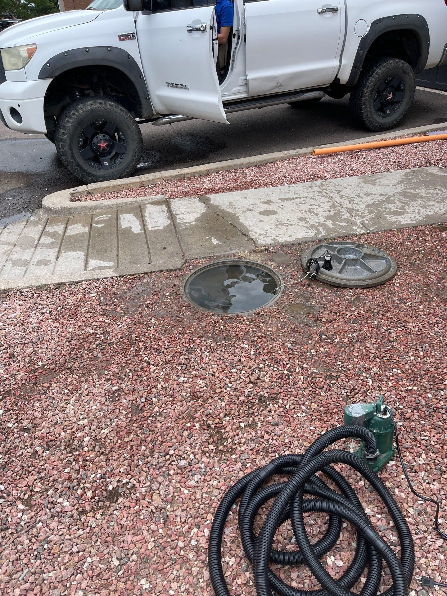White pickup truck with open door beside a wet sidewalk and utility covers, hose coiled in foreground