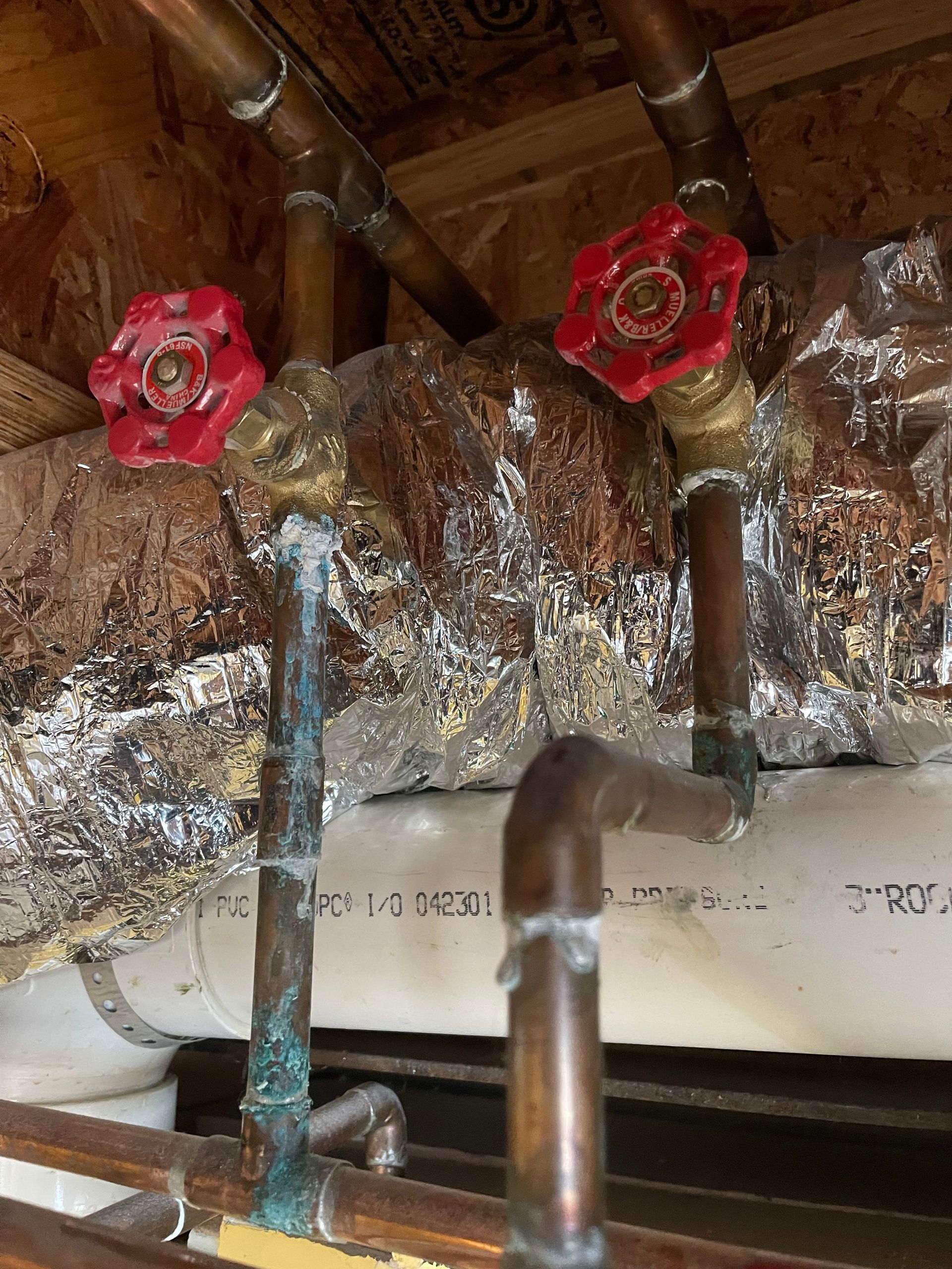 Red valve handles on copper pipes beneath insulated ducting in a crawlspace