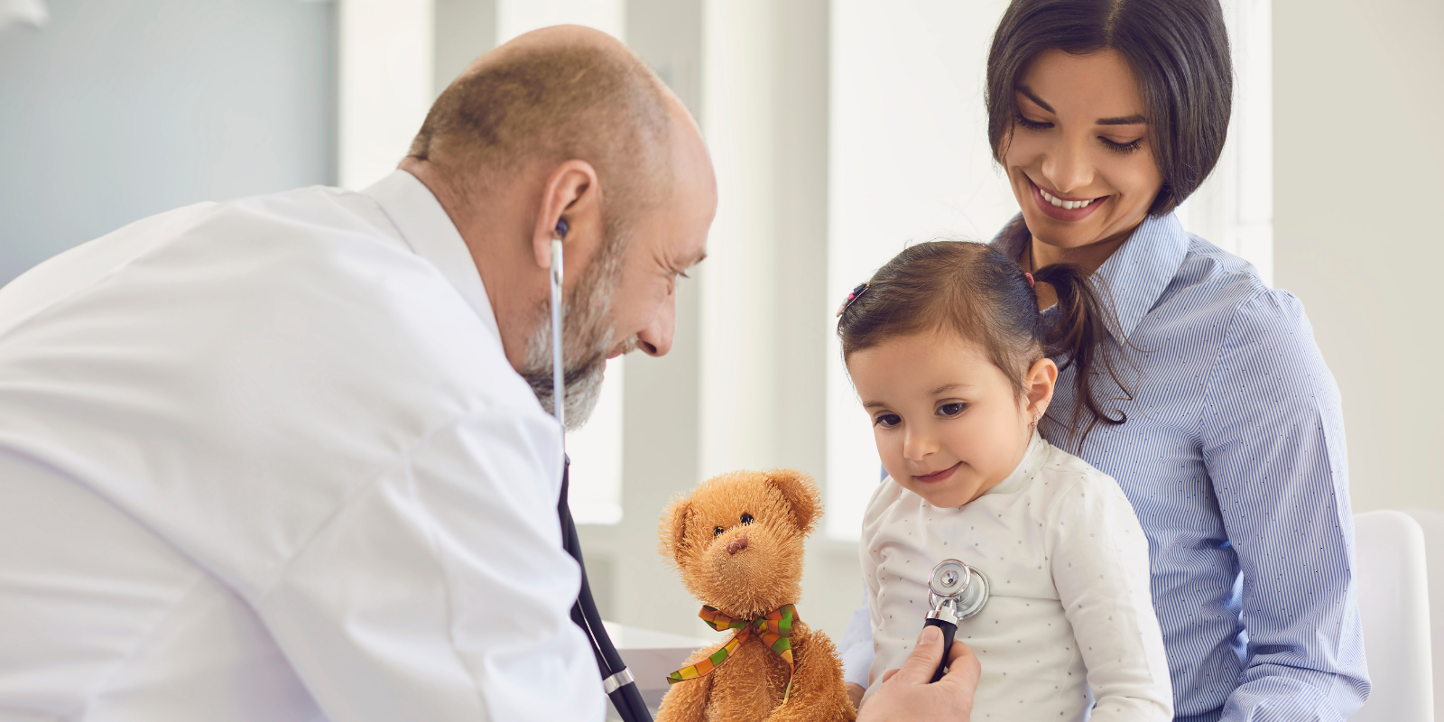Doctor examining a child with a teddy bear while a smiling parent stands nearby in a clinic