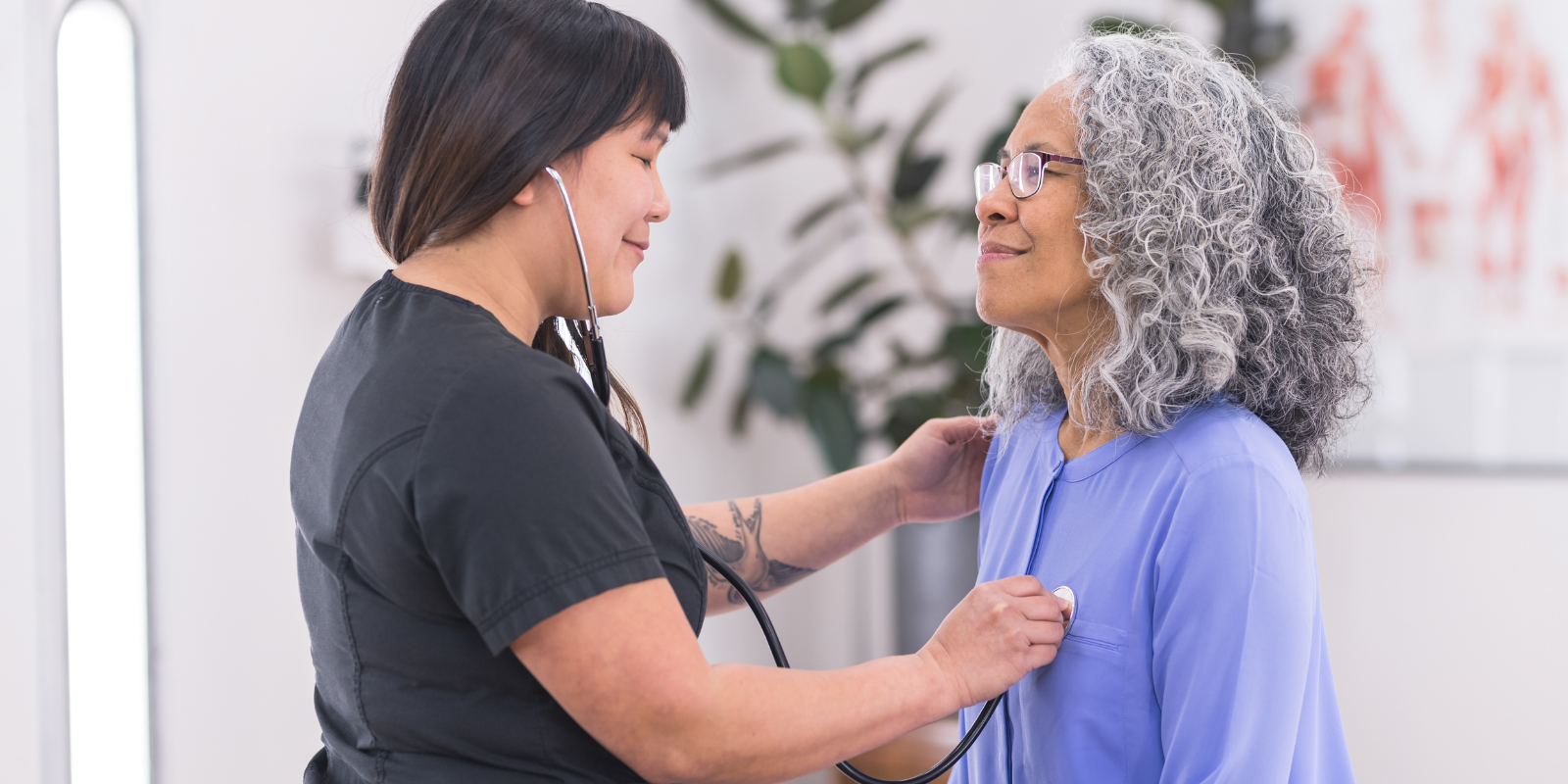 Two women smiling and talking indoors, one checking the other's medical gown or necklace.
