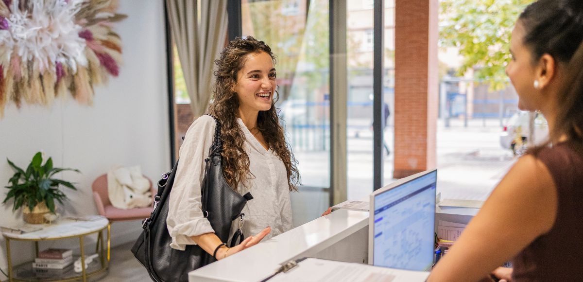 Two women chatting at a reception desk in a bright office lobby with a laptop open between them