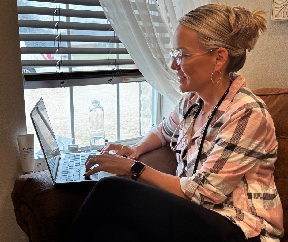 Woman working on laptop near a window, wearing glasses and a pink plaid shirt.
