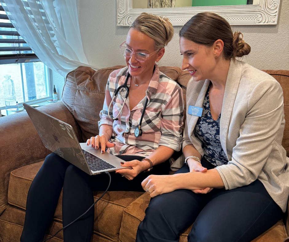 Two women looking at a laptop, smiling, on a brown couch. One wears a stethoscope.