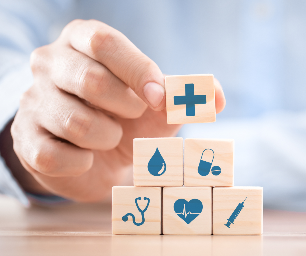 Hand placing a plus sign block atop stacked health-related icons: drop, pill, stethoscope, heart, syringe.