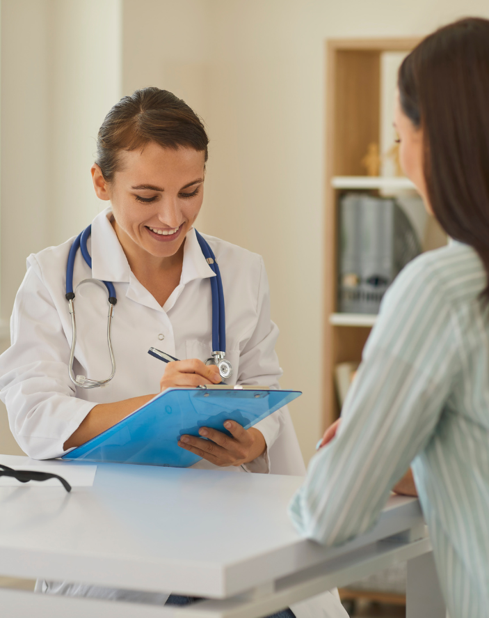 Doctor in a white coat with a stethoscope taking notes, smiling at a patient in an office setting.