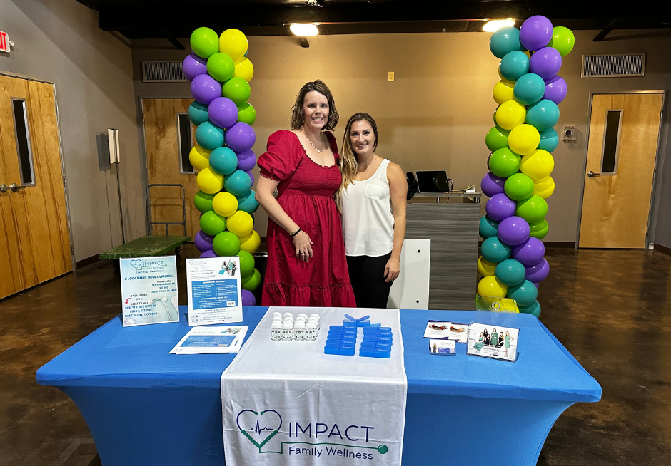 Two women stand behind a table at a health fair booth with balloon columns. The table has brochures and items for display.