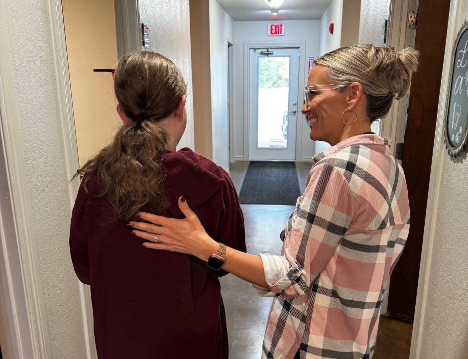 Woman with hand on another's back, hallway. Both smile, one in plaid shirt. Door with window in background.