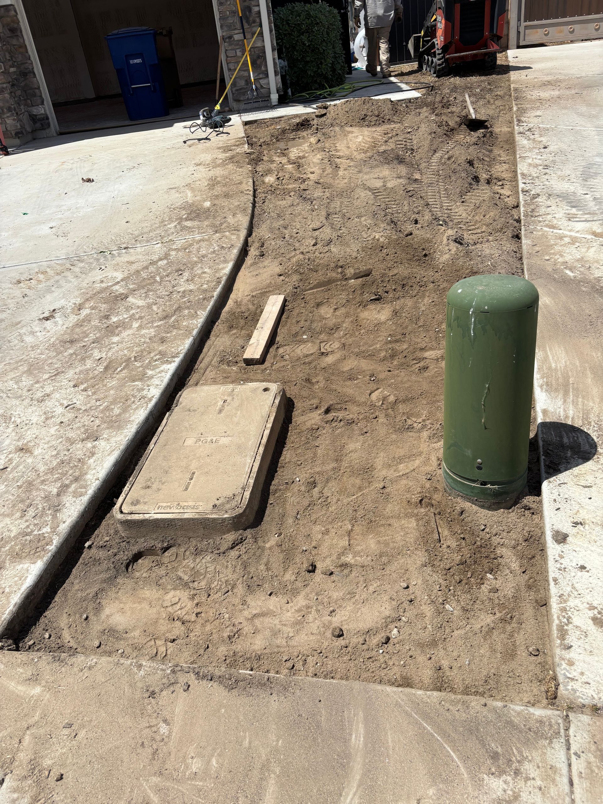 Utility trench and concrete sidewalk repair beside a green utility post on a dirt strip.