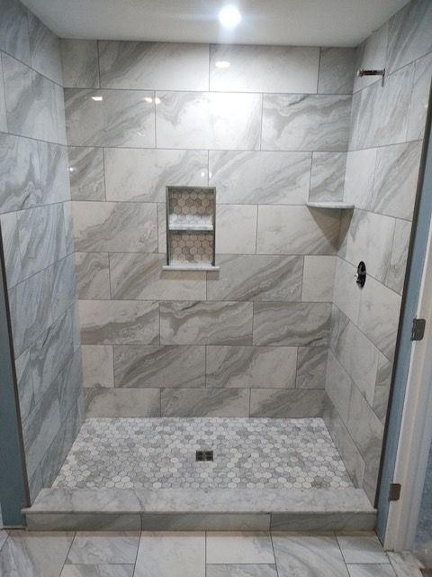 Empty tiled walk-in shower with marble-pattern walls, niche shelves, bench, and mosaic floor drain.