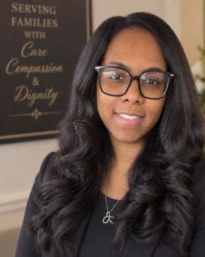 Woman with long dark hair and glasses smiling in an office, with a sign reading “Serving Families with Care Compassion and Dignity.”