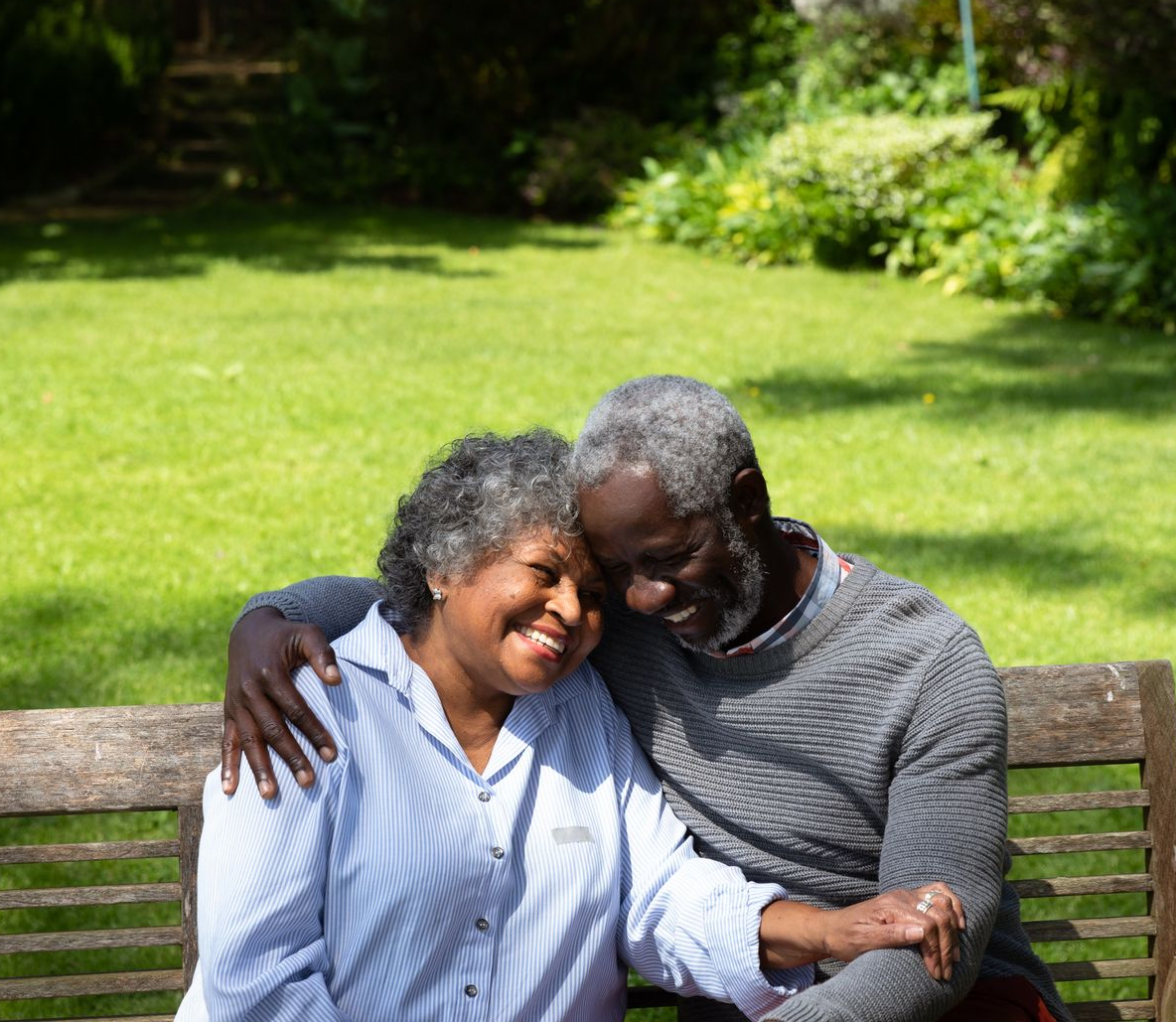 Couple smiling on a park bench, leaning into each other in a sunlit green garden.