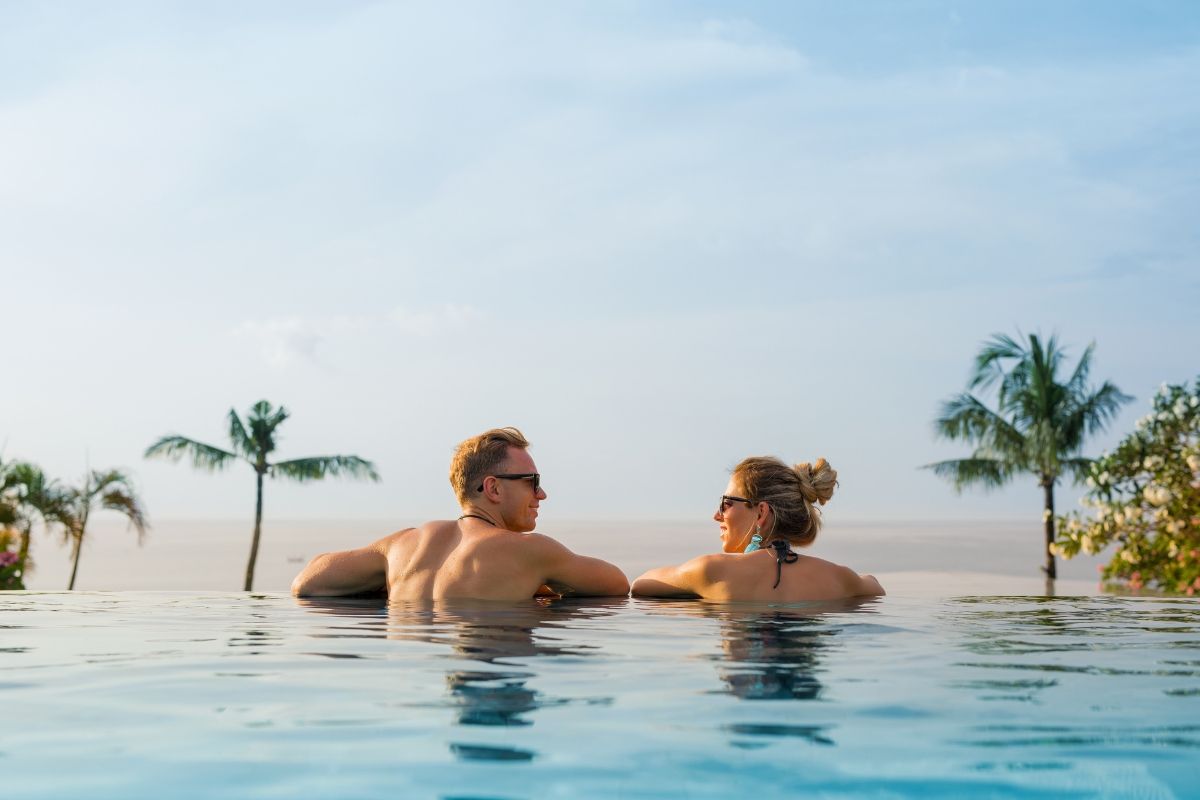Casal em uma piscina infinita, olhando um para o outro, em um cenário tropical com palmeiras e vista para o mar.