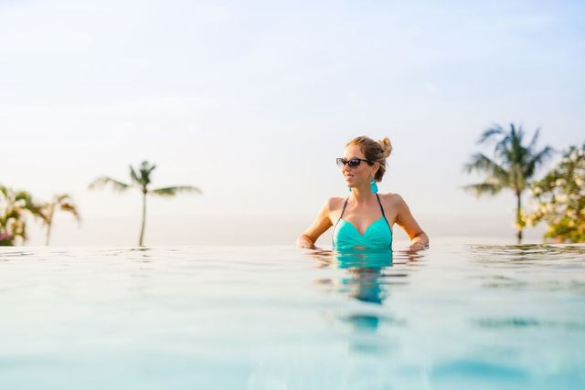 Mulher de maiô turquesa relaxando em uma piscina infinita tropical com palmeiras ao fundo.