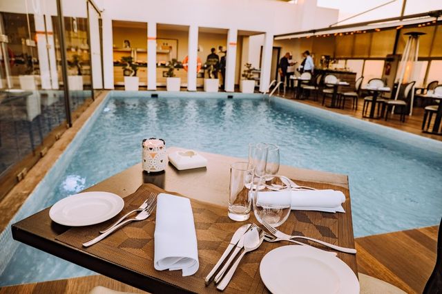 Mesa de jantar ao lado de uma piscina coberta, posta com pratos brancos e taças de vinho em um ambiente de resort.