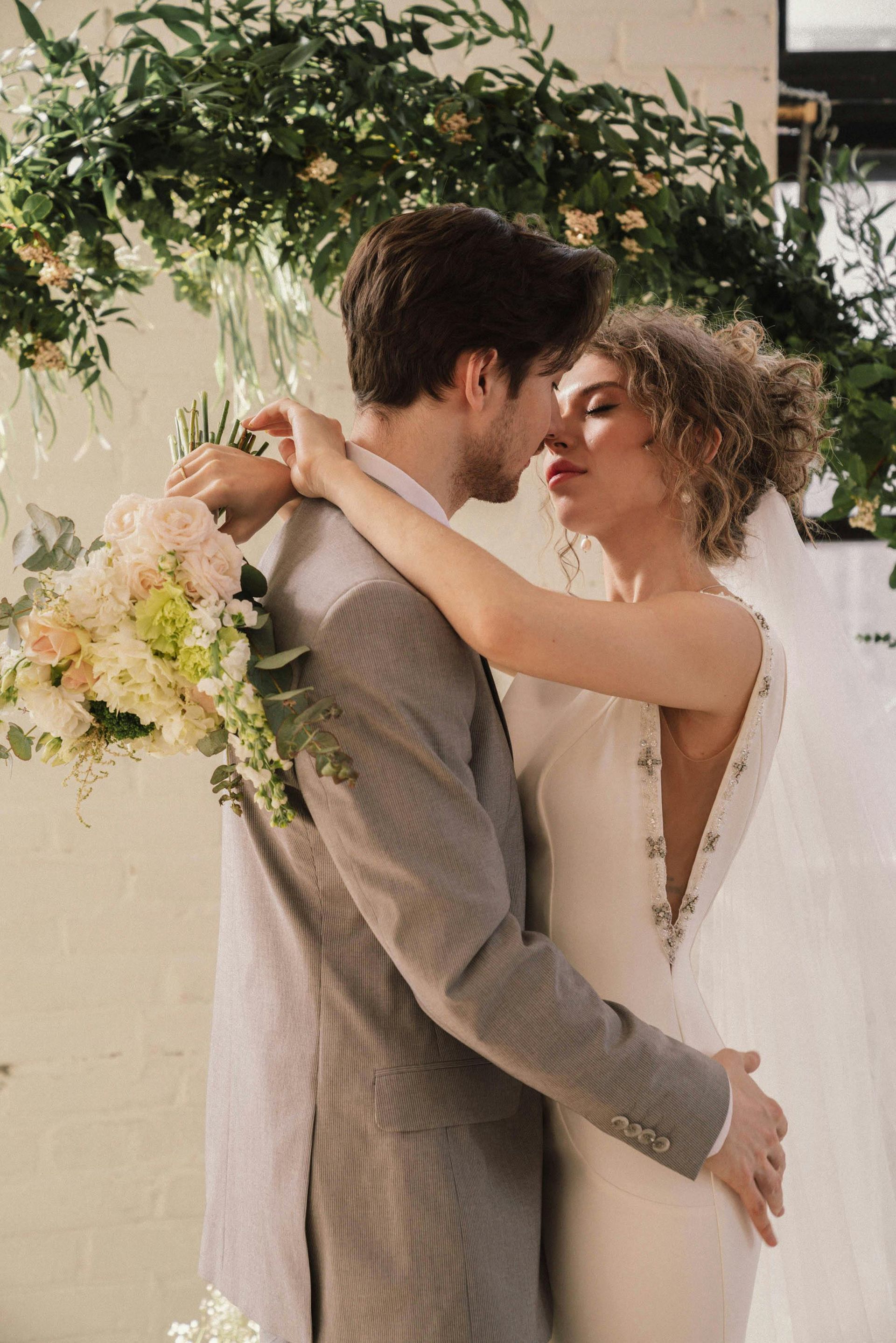 Casal se abraçando na cerimônia de casamento, com folhagens e um buquê ao fundo.