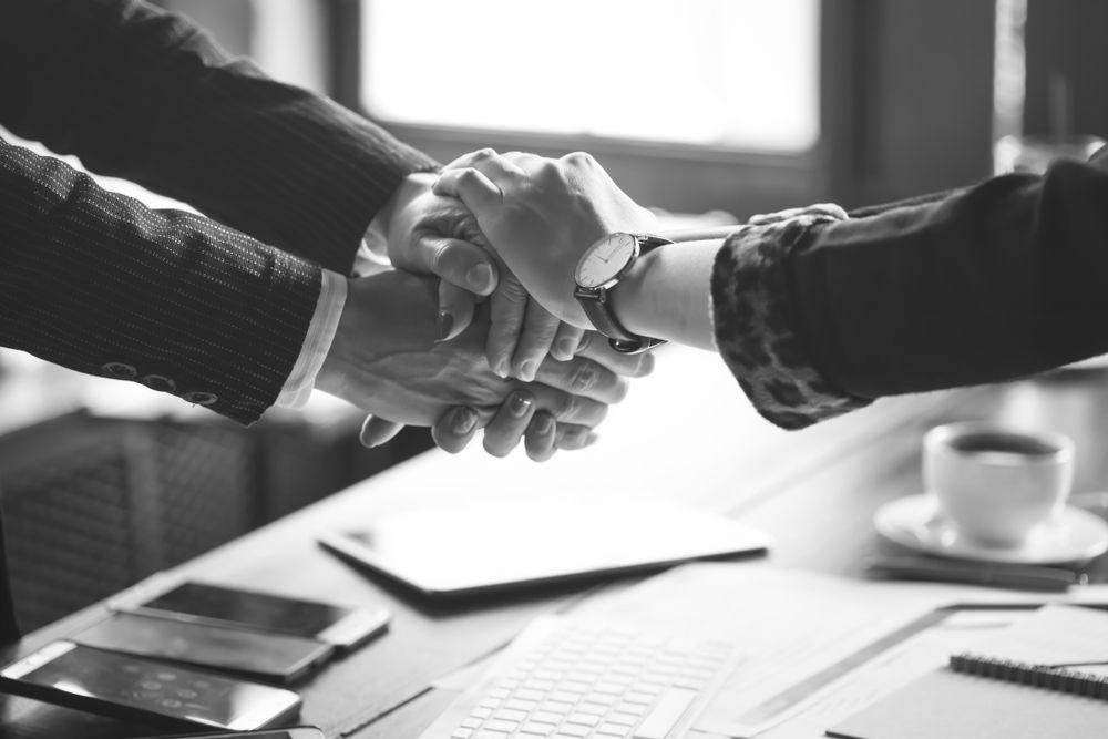 Two people shake hands across a desk with papers, a laptop, and a coffee cup in black-and-white. - ev law in Bundall, QLD