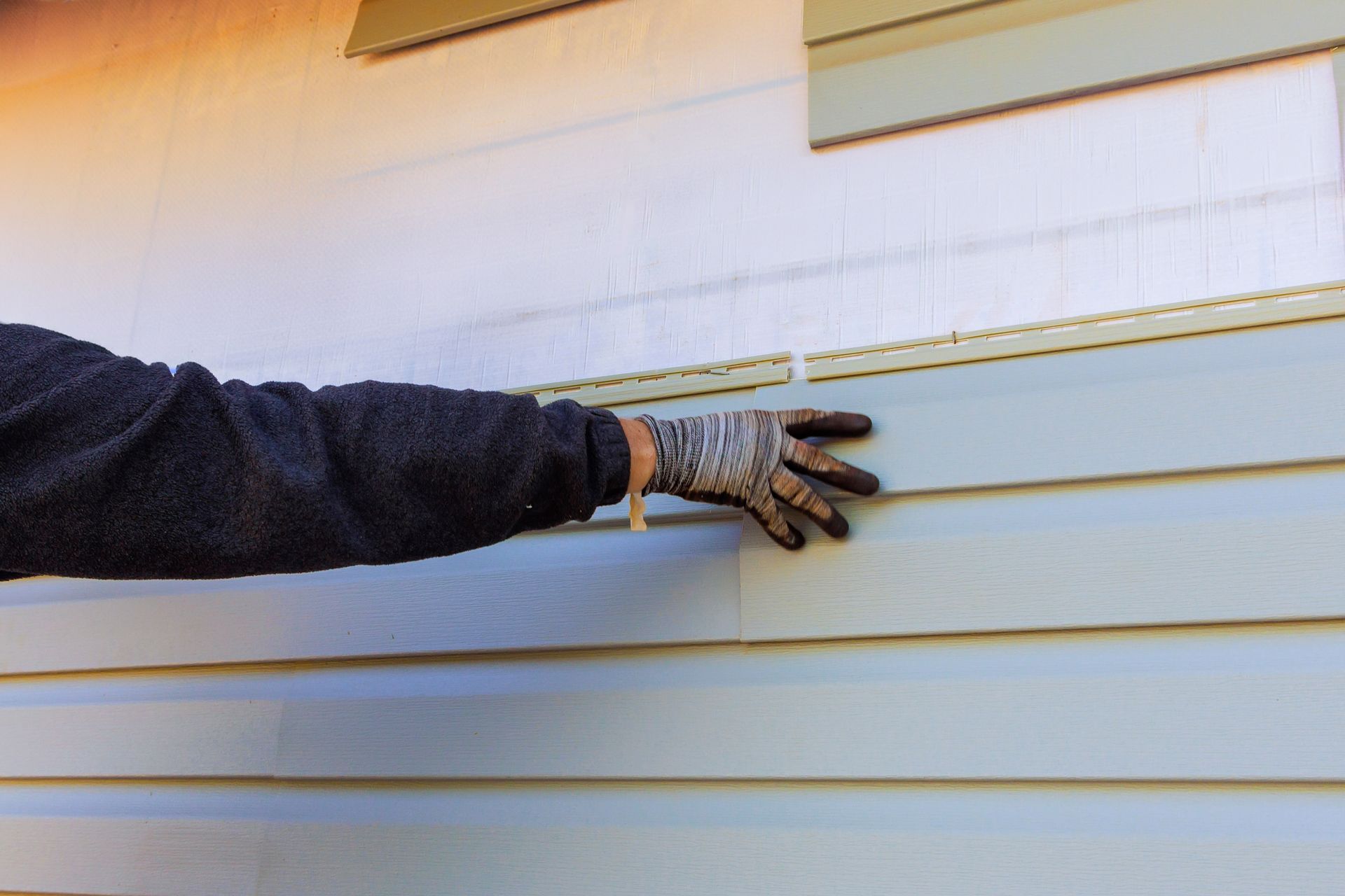 Hand with work glove touching light blue siding on a building.