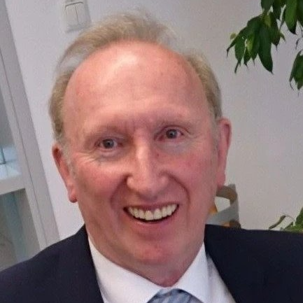 Ein lächelnder Mann in dunklem Anzug und weißem Hemd in einem Innenraum, im Hintergrund eine Pflanze.