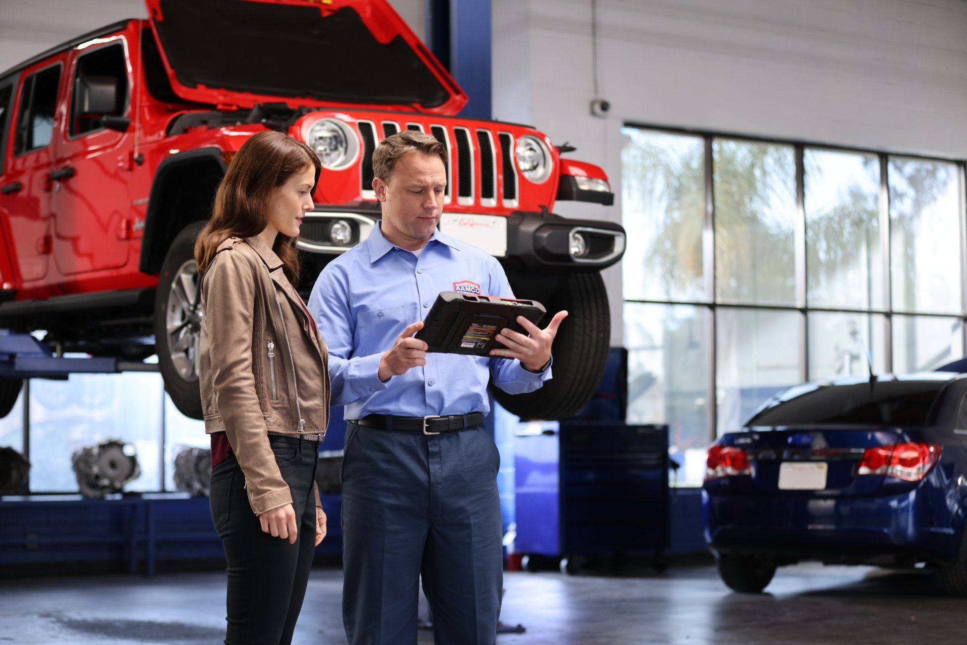 Mechanic showing a woman a tablet in a garage with a red car raised on a lift.