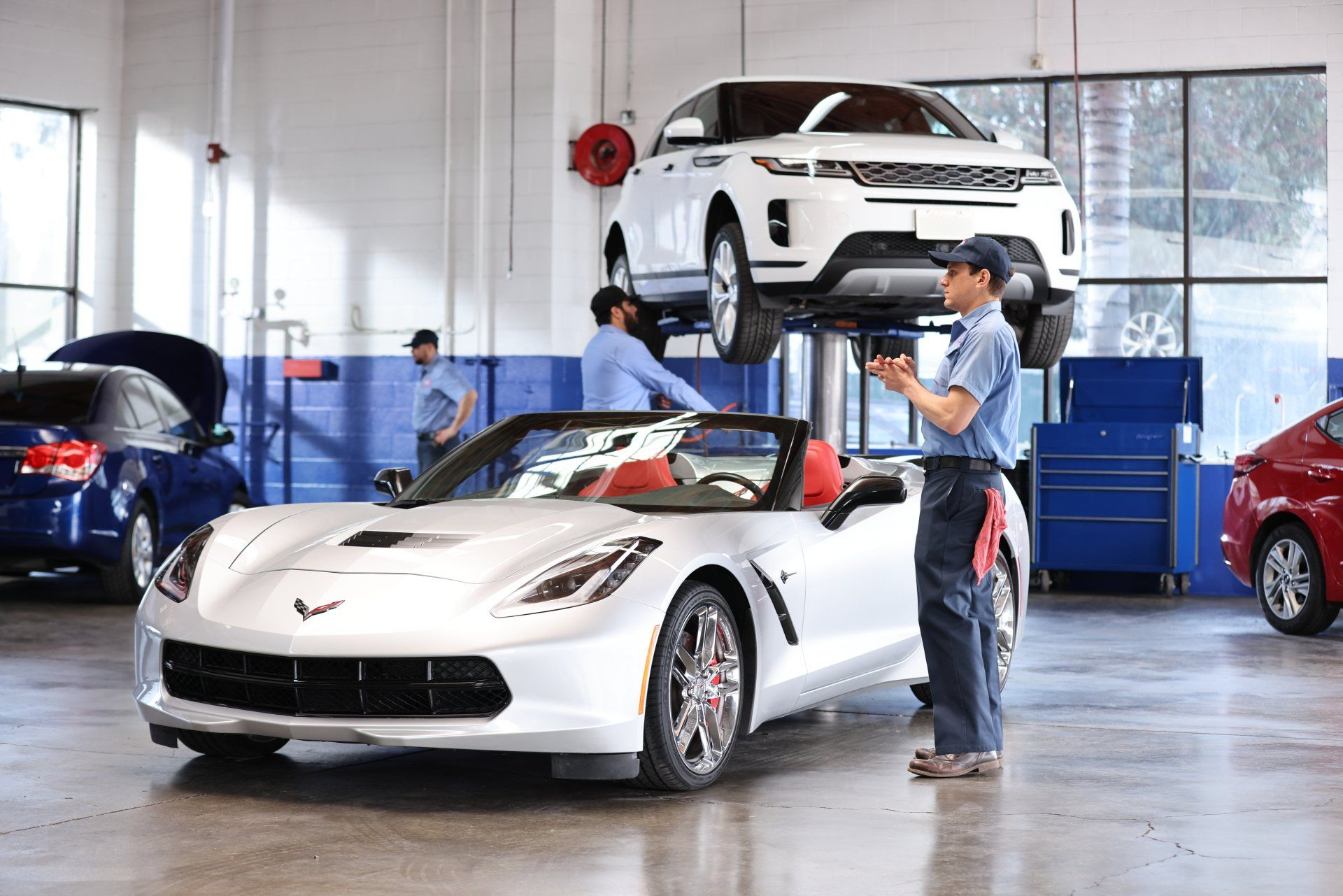 Mechanics in a car repair shop. A white convertible and an SUV on a lift. Other cars visible.