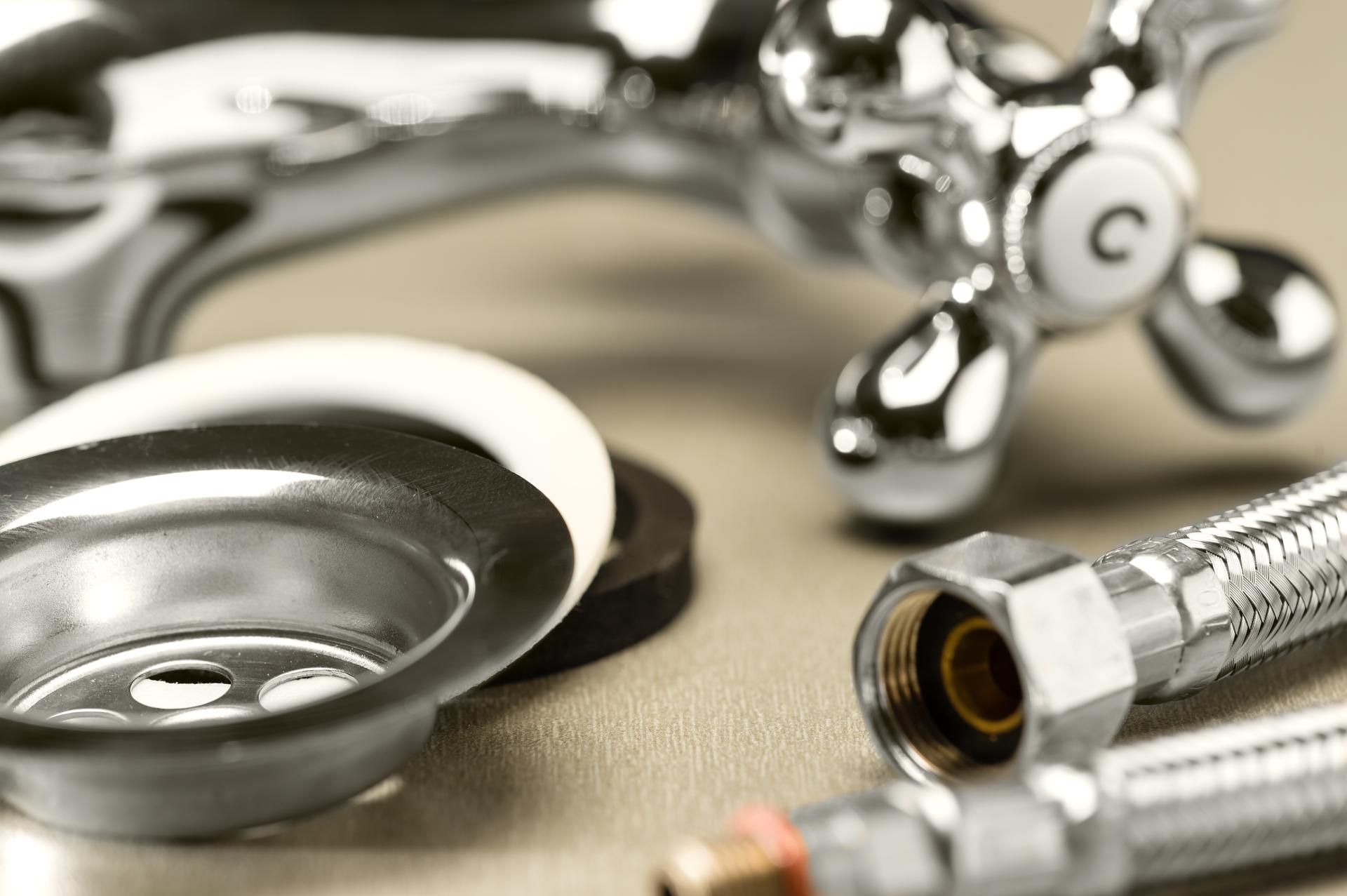 A chrome faucet handle, a sink drain assembly with gaskets, and flexible supply lines arranged on a neutral surface.