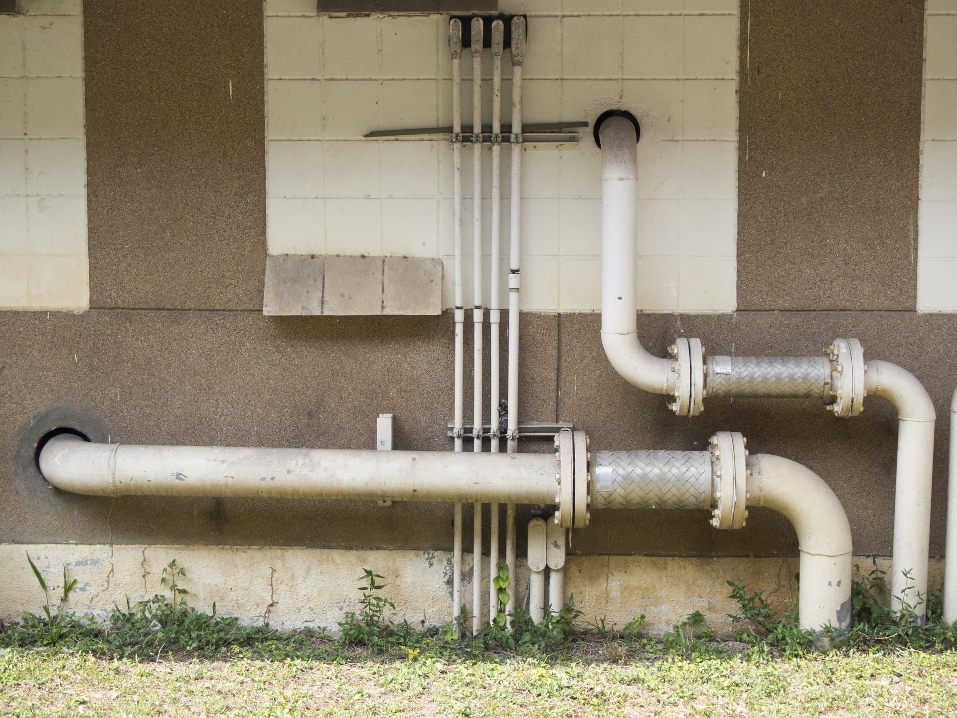 Two white industrial pipes with flanged connectors run horizontally and vertically along a beige and brown exterior wall.