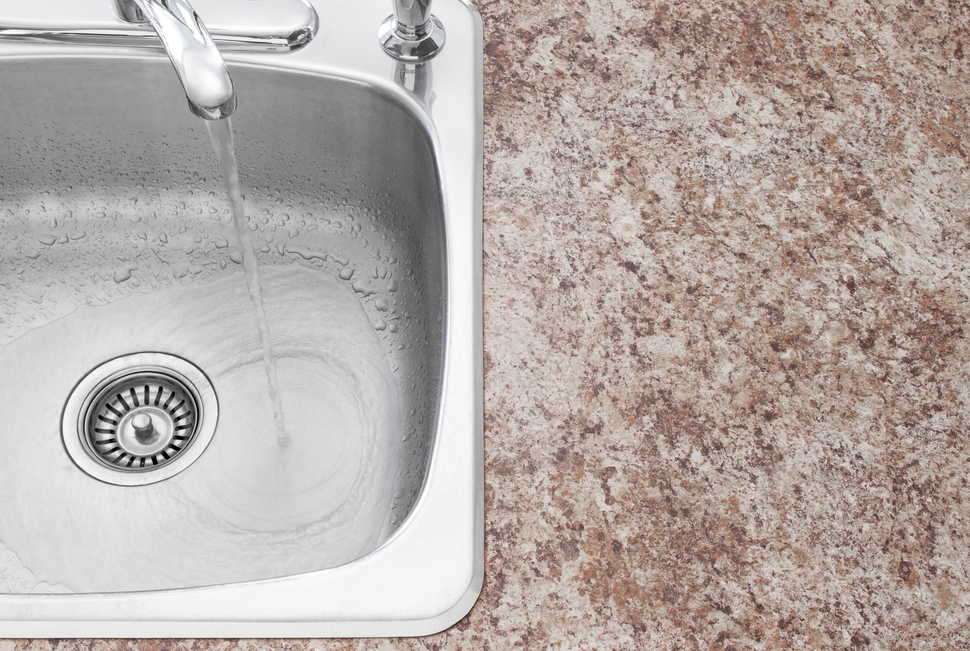 A silver kitchen faucet pours water into a stainless steel sink beside a textured, brown-toned countertop.