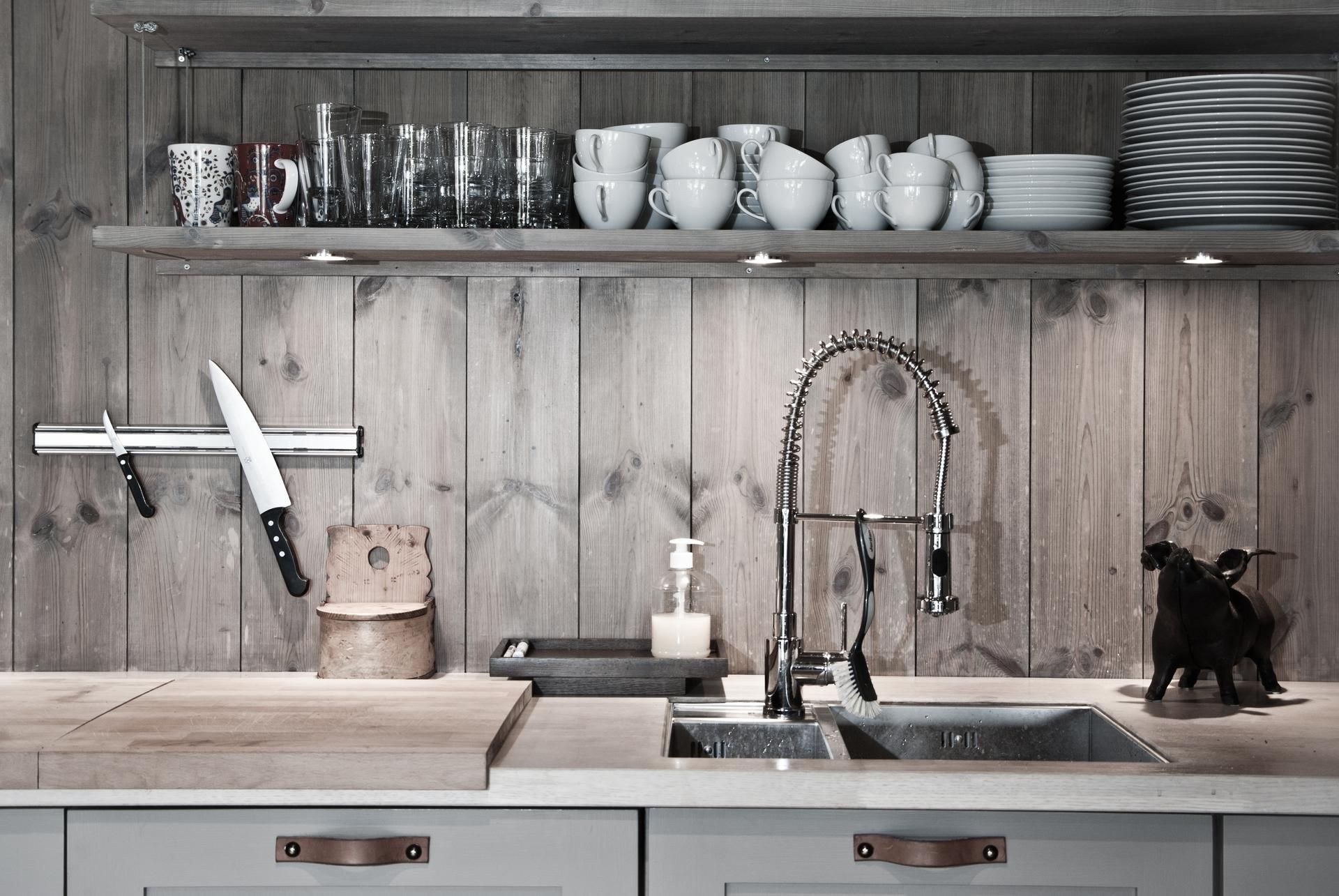 A rustic kitchen sink area with a stainless steel faucet, wooden wall paneling, a knife rack, and an open storage shelf.