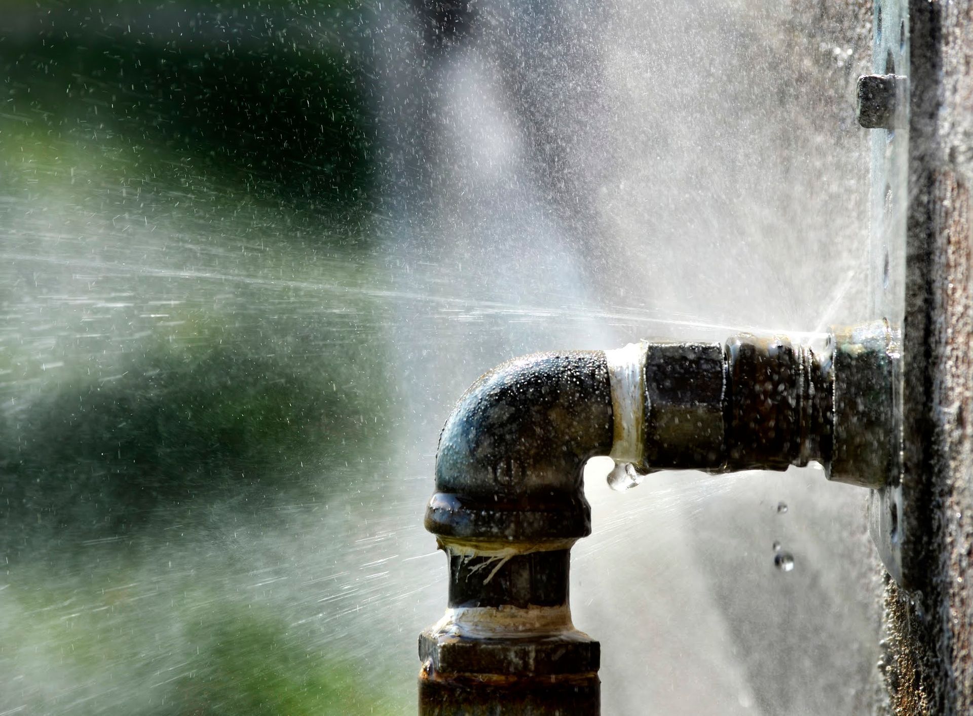 A metal plumbing elbow joint attached to a wall, spraying a fine mist of water from a leak at the threaded connection.