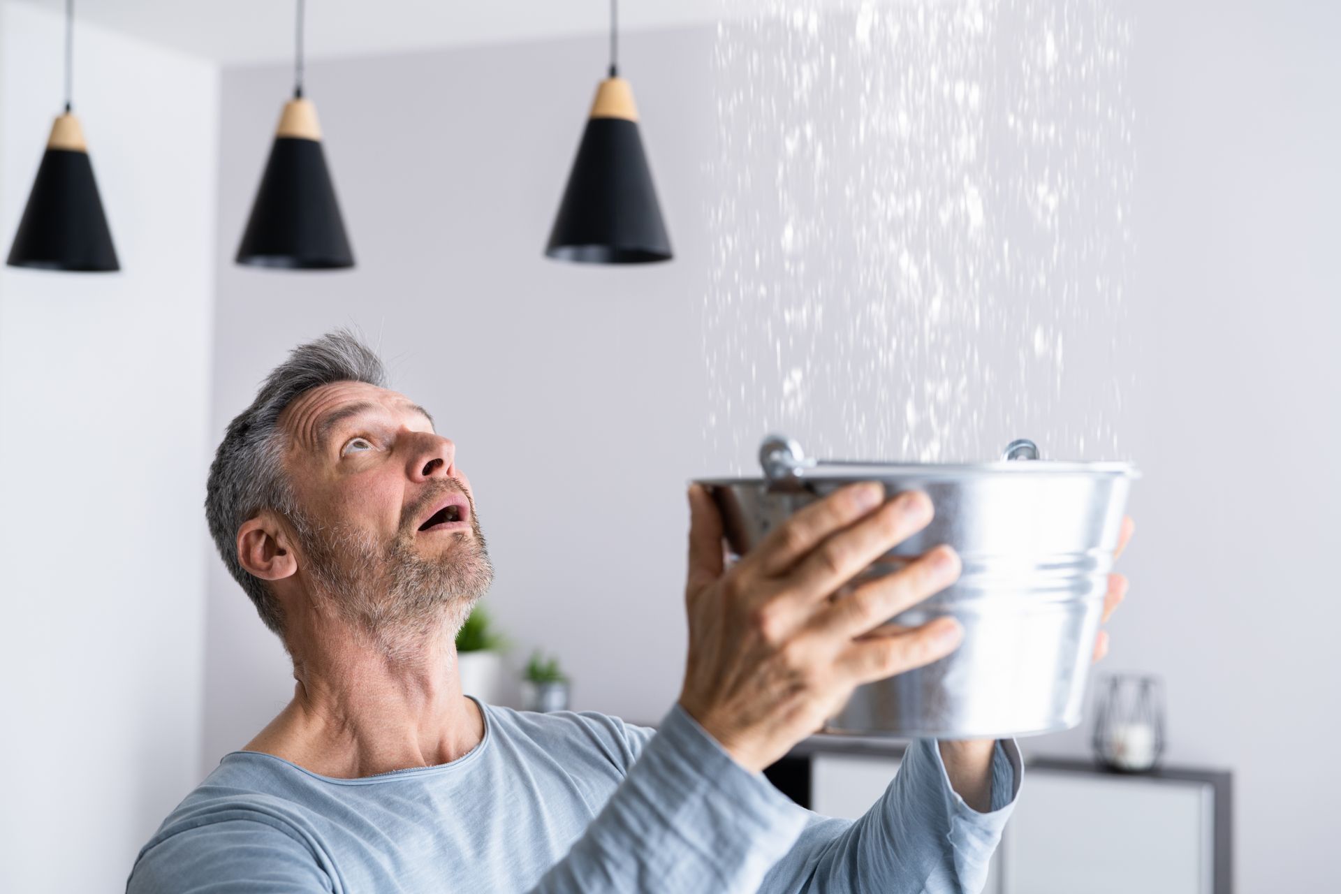 Person holding a metal bucket under a heavy ceiling leak caused by a burst pipe.